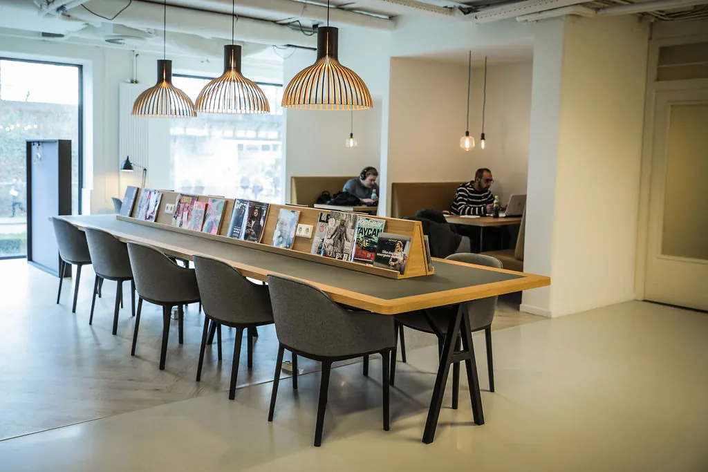 Moderne, open werkruimte aan de Marten Meesweg met een lange leestafel, tijdschriften en mensen aan het werk op laptops.